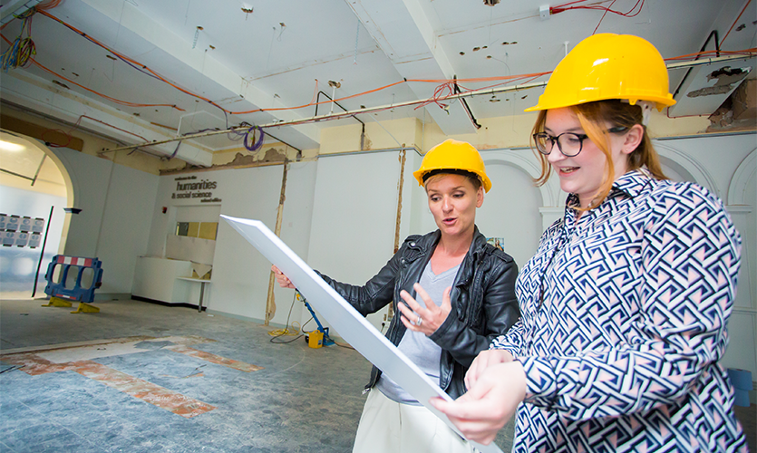 Two women wearing hard hats looking at plans inside a room under construction